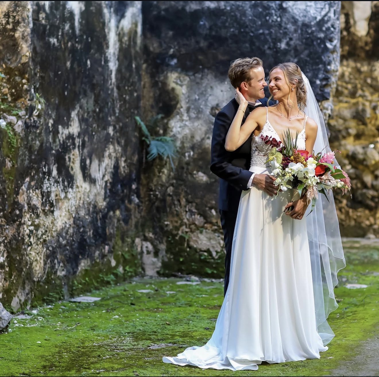Novios abrazados en un jardín de hacienda con muros de piedra