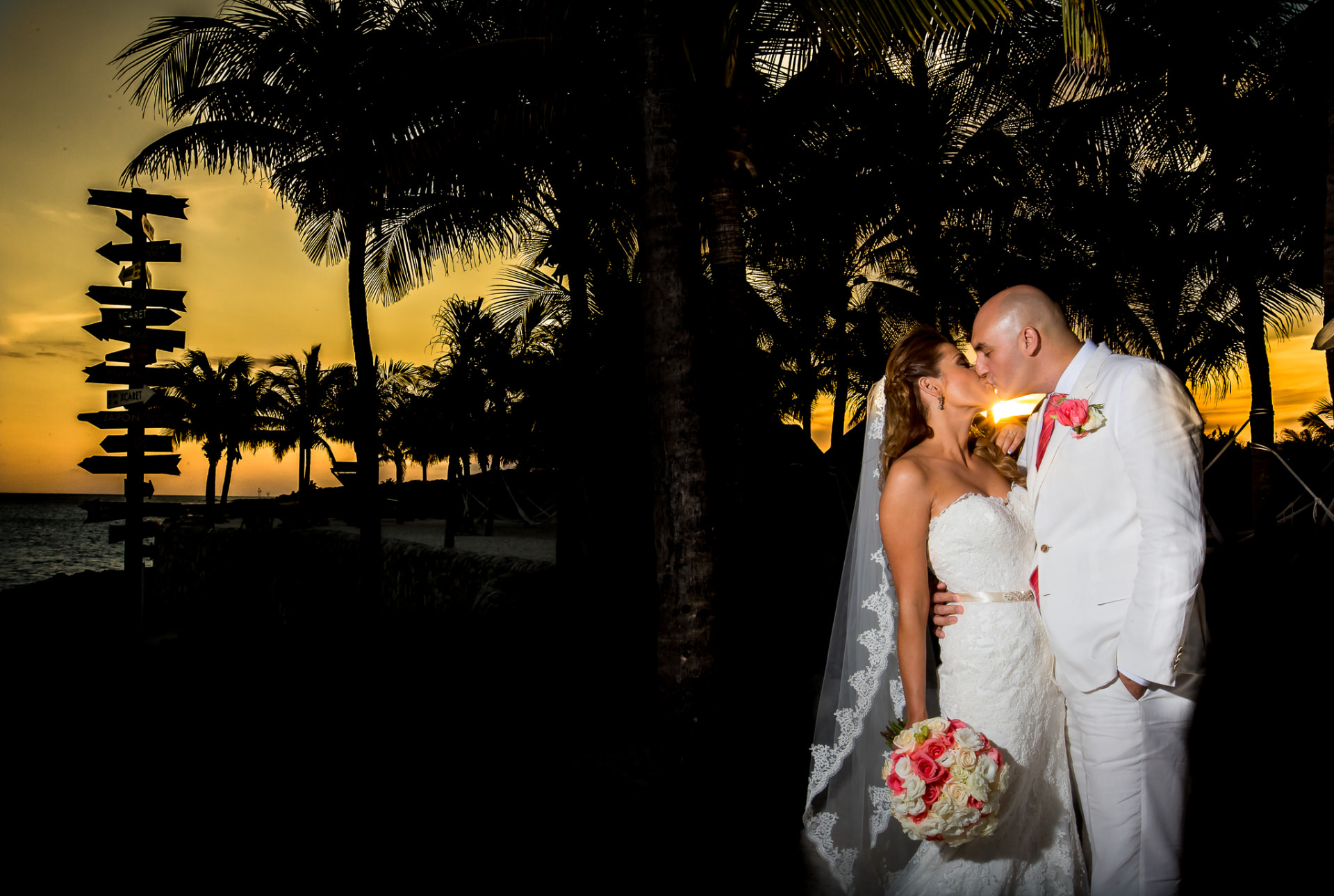 Novios besándose al atardecer entre palmeras frente al mar